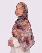 A woman wearing a vibrant printed modal hijab with an intricate multi-color marble pattern featuring orange, purple, and charcoal grey tones, paired with a cream outfit