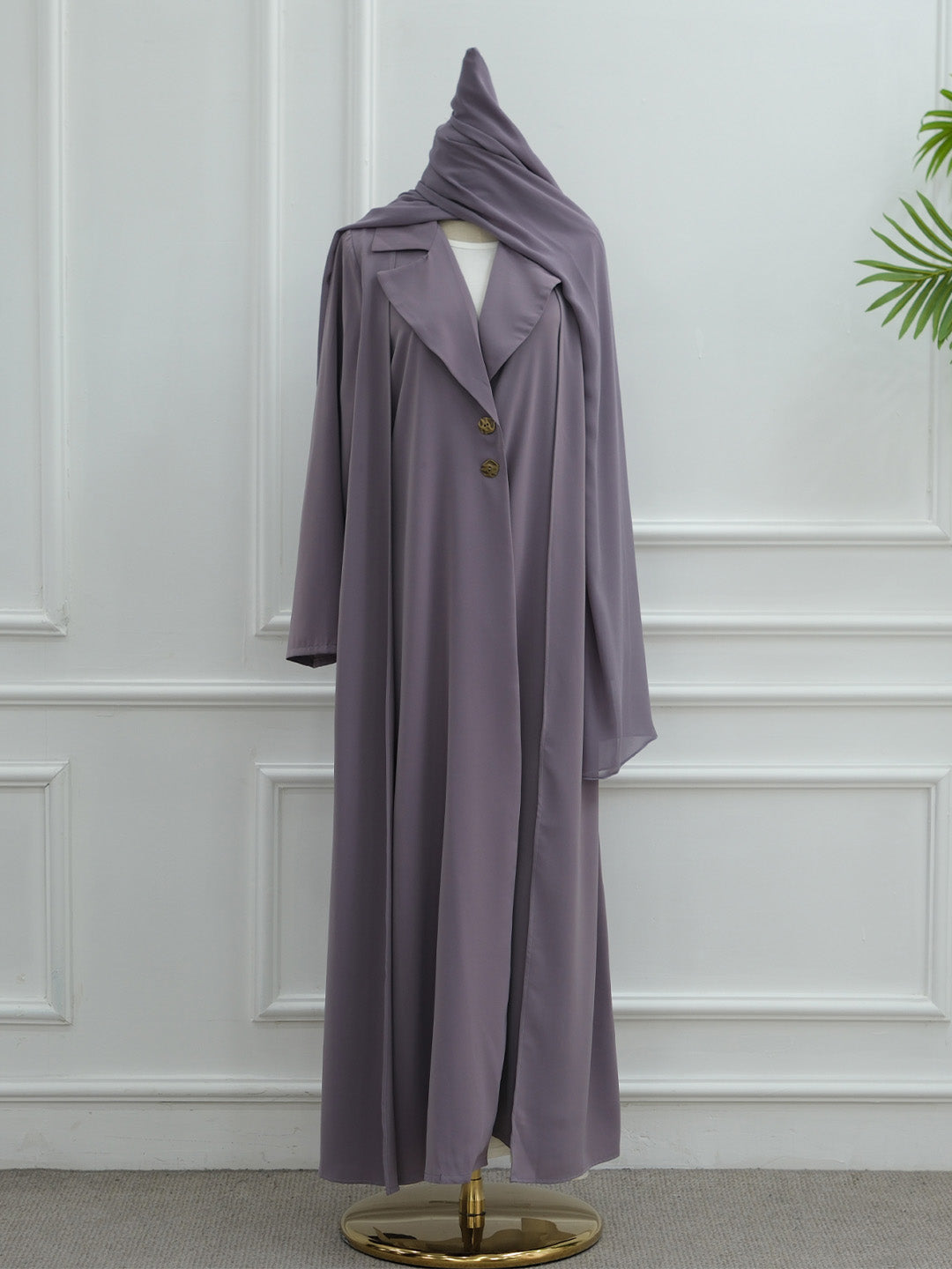 A long, flowing abaya dress in a muted dusty lavender-purple color, displayed on a mannequin with a matching draped headscarf. The abaya features a single row of decorative gold buttons down the front.