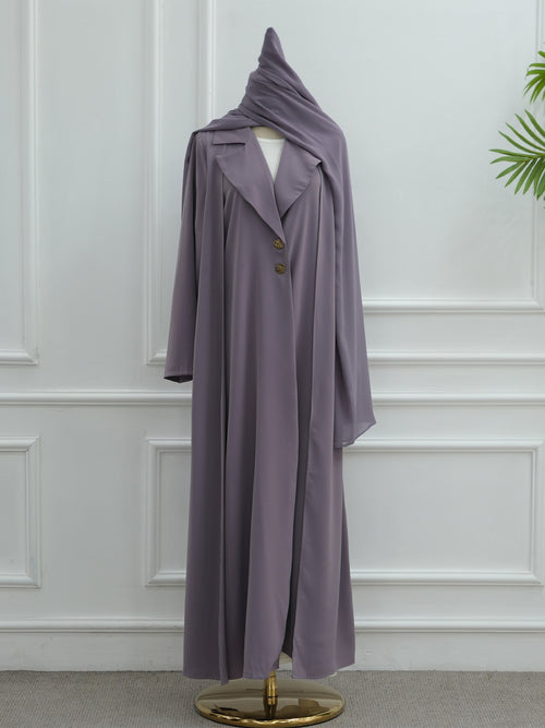A long, flowing abaya dress in a muted dusty lavender-purple color, displayed on a mannequin with a matching draped headscarf. The abaya features a single row of decorative gold buttons down the front.