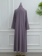 A long, loose-fitting abaya dress in a muted dusty lavender-purple color, shown from the back on a mannequin with a matching pointed head covering. The mannequin is set against a white paneled wall with a green plant leaf visible in the upper right corner.