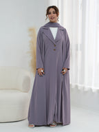 A woman models a long, flowing mauve-purple abaya dress with a matching headscarf, worn over a white top. The dress features a double-breasted front with brown buttons and a relaxed silhouette, styled in a bright, modern room with a white curved sofa and sheer curtains.