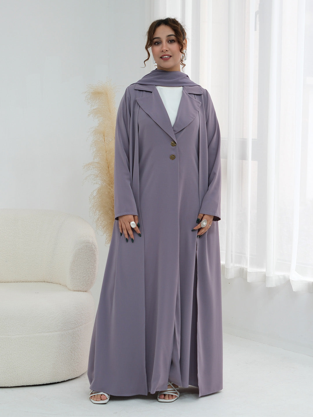 A woman models a long, flowing mauve-purple abaya dress with a matching headscarf, worn over a white top. The dress features a double-breasted front with brown buttons and a relaxed silhouette, styled in a bright, modern room with a white curved sofa and sheer curtains.