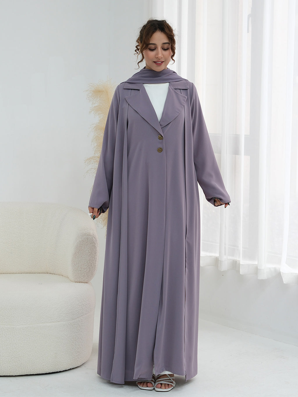 A woman modeling a long, flowing mauve-purple abaya dress with a matching head scarf, worn over a white top. The abaya features a notched lapel, three front buttons, and wide sleeves, styled in a bright, modern room with a white curved sofa and sheer curtains.