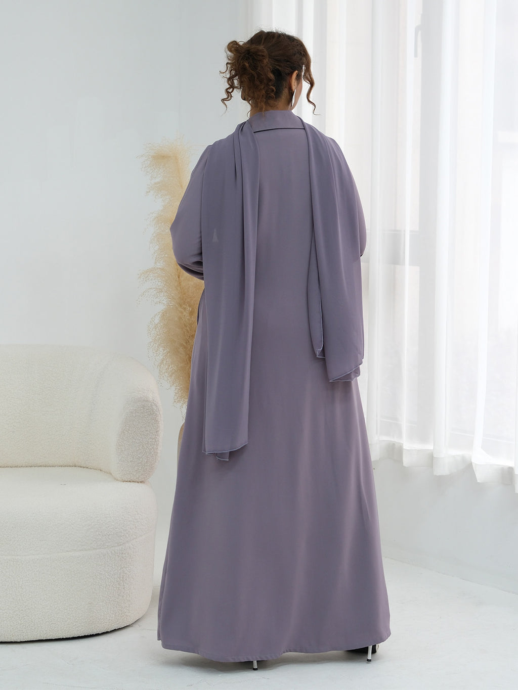 A woman viewed from behind wearing a long, flowing abaya in a muted dusty lavender-purple color, standing in a bright, minimalist room with white curtains and a cream-colored armchair.