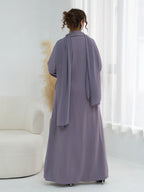 A woman viewed from behind wearing a long, flowing abaya in a muted dusty lavender-purple color, standing in a bright, minimalist room with white curtains and a cream-colored armchair.