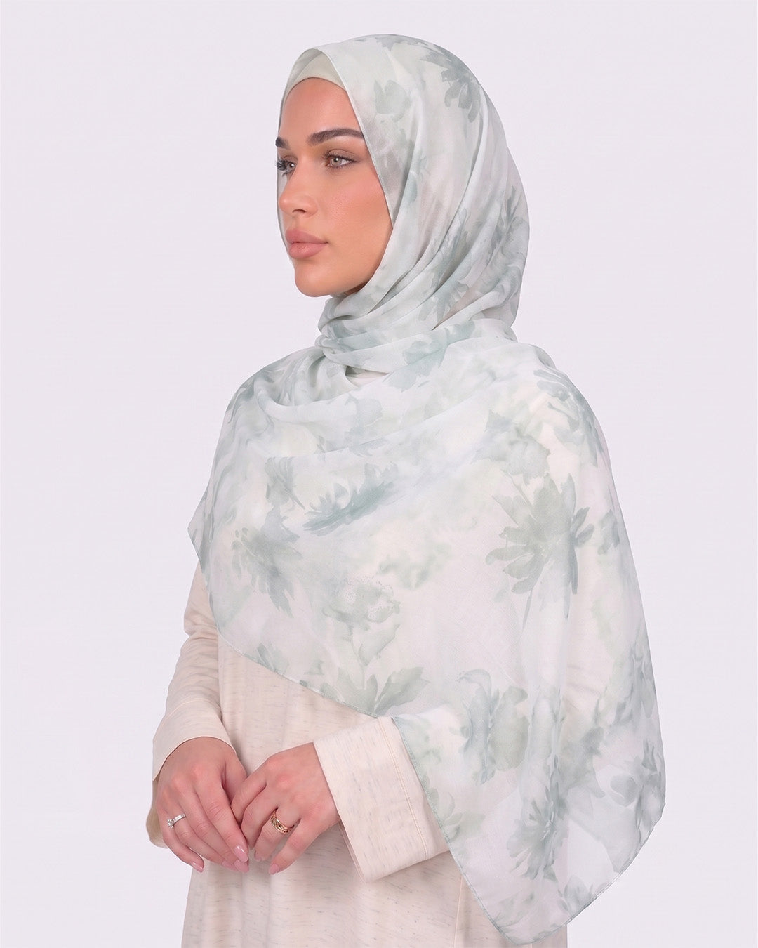 A woman wearing a fresh white printed modal hijab with soft sage and mint green watercolor floral motifs, paired with a cream outfit.