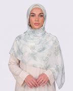 A woman wearing a fresh white printed modal hijab with soft sage and mint green watercolor floral motifs, paired with a cream outfit.