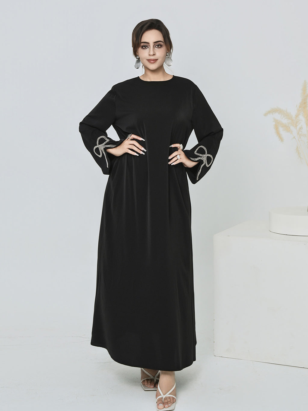 A woman modeling a long black dress with wide sleeves and decorative white bow details at the cuffs, standing against a light studio background.
