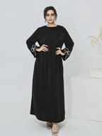 A woman modeling a long black dress with wide sleeves and decorative white bow details at the cuffs, standing against a light studio background.