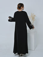 A woman wearing a long black abaya dress with silver bow details on the sleeves, shown from the back in a studio setting with a white pedestal and dried pampas grass.