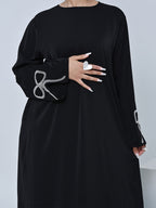 A black long-sleeve dress with a decorative silver rhinestone bow on the right sleeve, shown on a model against a light gray background.