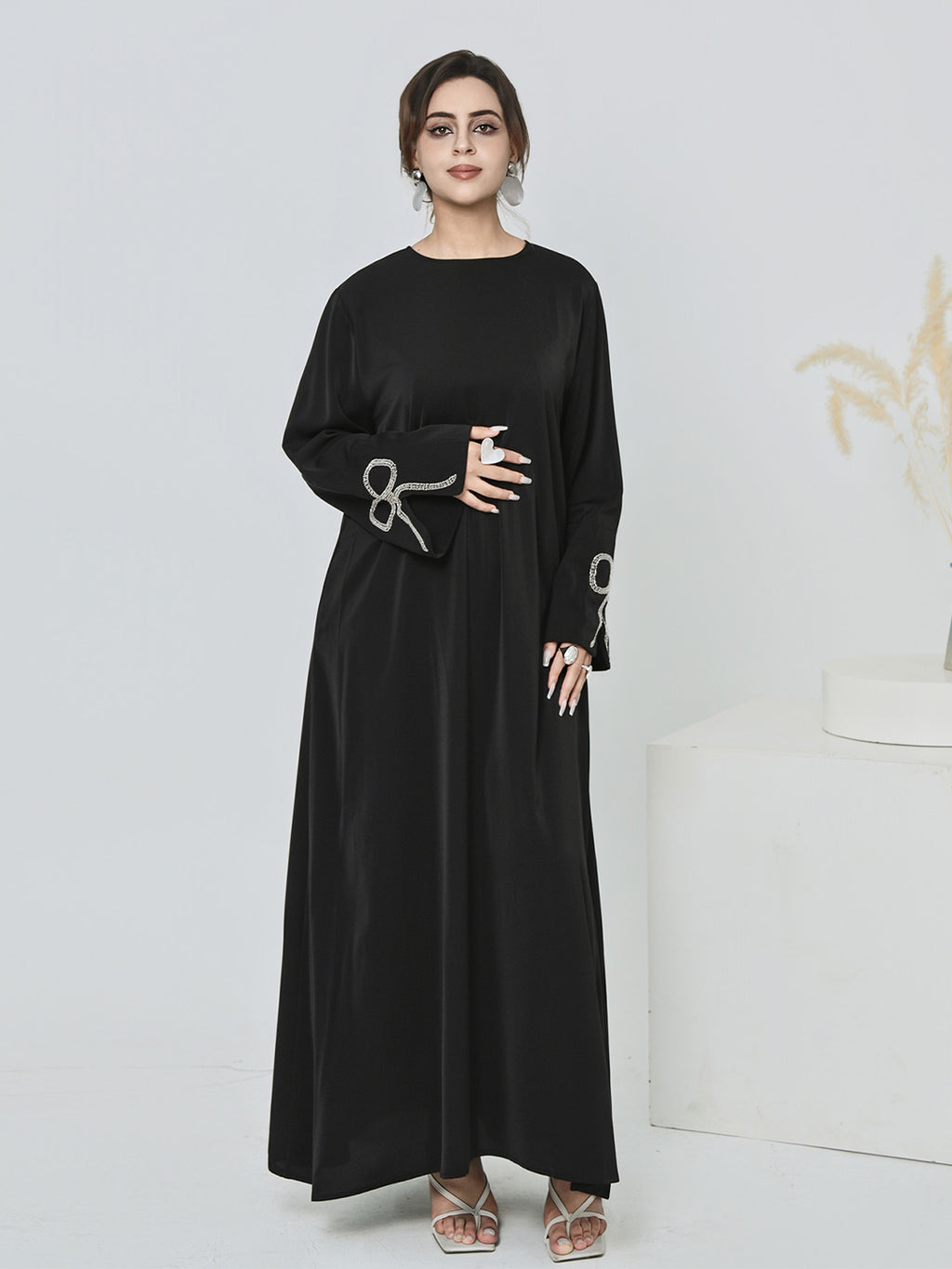 A woman modeling a long black abaya dress with wide sleeves and silver decorative embroidery on the cuffs, standing against a clean white studio background.