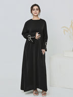 A woman modeling a long black abaya dress with wide sleeves and silver decorative embroidery on the cuffs, standing against a clean white studio background.