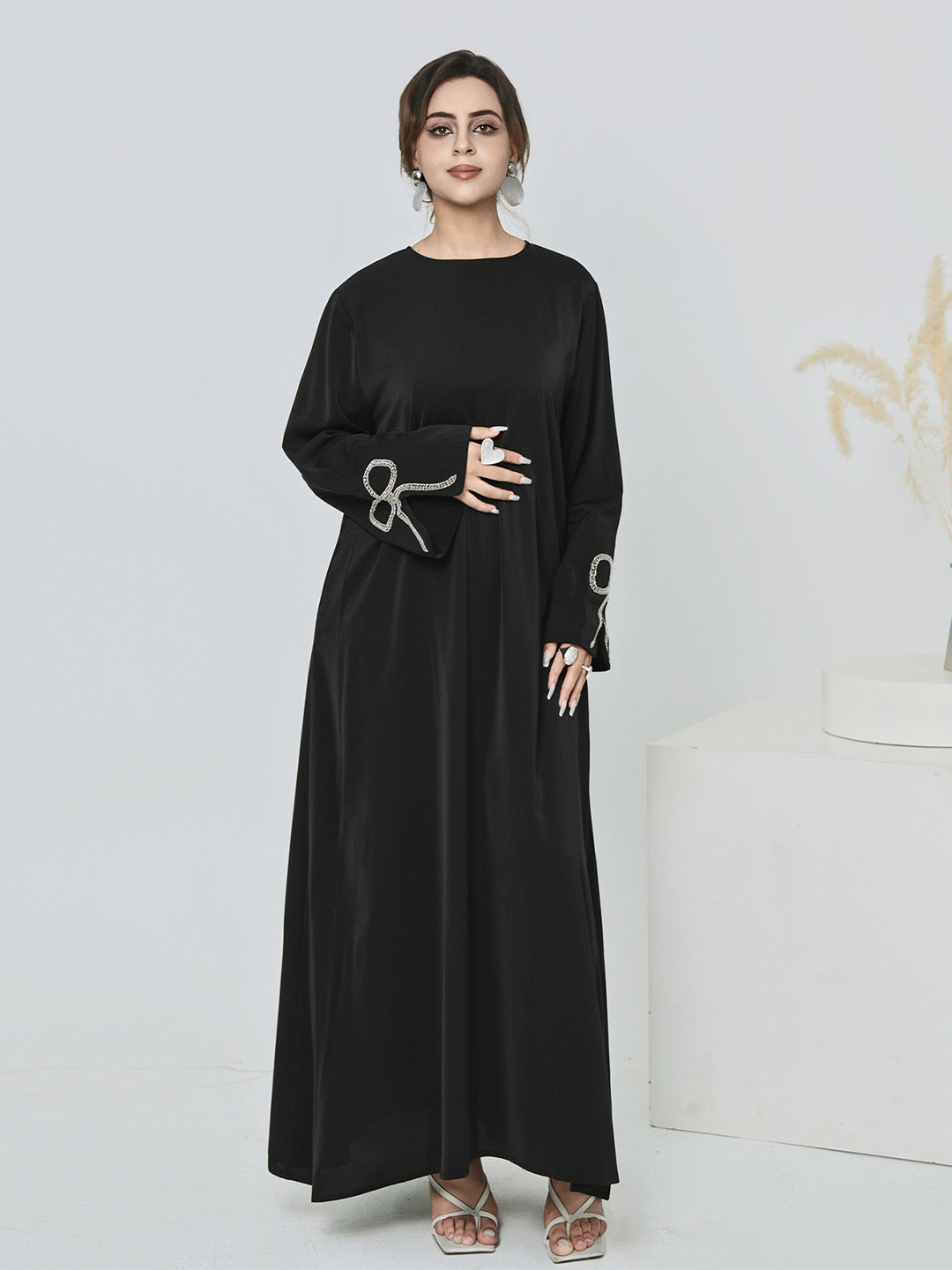 A woman modeling a long black abaya dress with wide sleeves and silver decorative embroidery on the cuffs, standing against a clean white studio background.