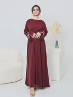 A woman modeling a long-sleeved, floor-length maroon dress with white embroidered details on the cuffs, standing in a bright, modern studio setting with a white sofa and decorative pedestal.