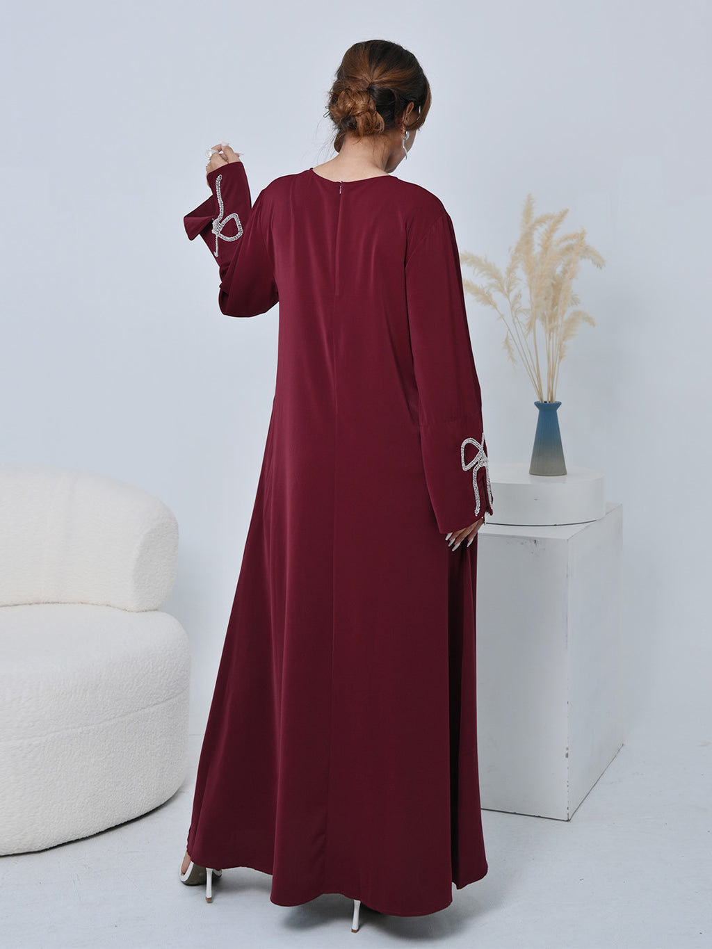 A woman wearing a long-sleeved maroon maxi dress, shown from the back in a studio setting with a white armchair and a decorative pedestal in the background.