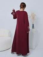 A woman wearing a long-sleeved maroon maxi dress, shown from the back in a studio setting with a white armchair and a decorative pedestal in the background.