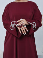 A close-up of a woman wearing a long-sleeved burgundy dress with silver rhinestone bow embellishments on the cuffs, shown from the shoulders to the hips against a light gray studio background.