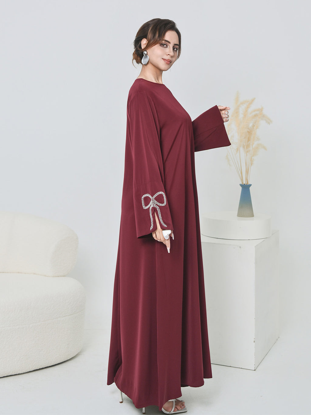 A woman modeling a long-sleeved, floor-length maroon abaya dress with white embroidered details on the sleeve, standing in a bright, modern studio setting.