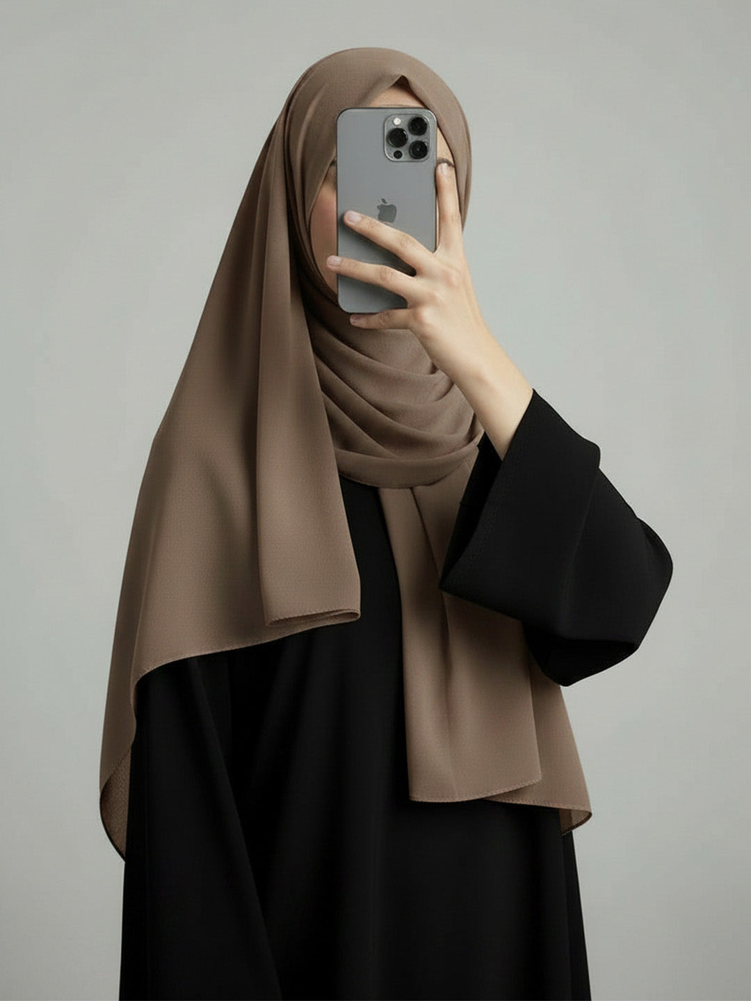 A woman wearing a taupe-brown hijab and a black abaya takes a mirror selfie with her phone against a plain gray background.