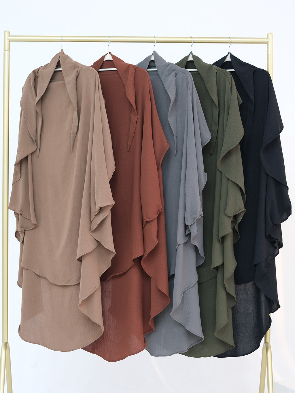 Five lightweight draped shawls or kaftan-style tops in different colors hang on a gold clothing rack against a white background. The colors shown are taupe, terracotta, slate blue, olive green, and black.