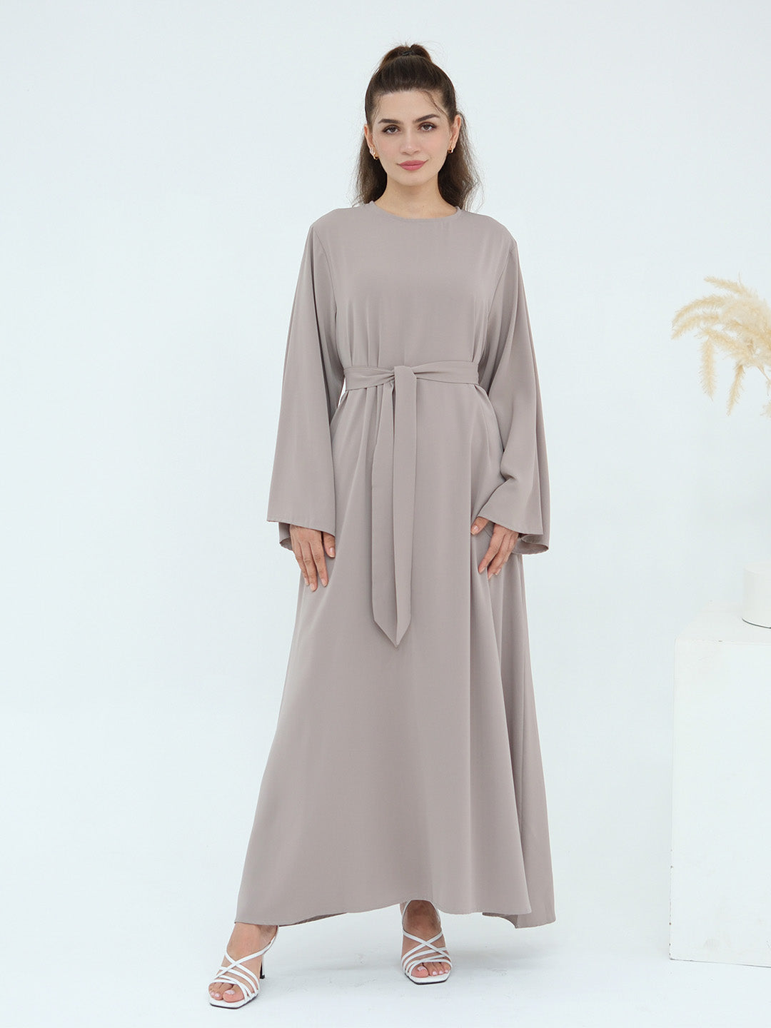 A woman modeling a long-sleeved, floor-length taupe maxi dress with a matching waist tie, paired with white strappy heels, set against a clean white studio background.