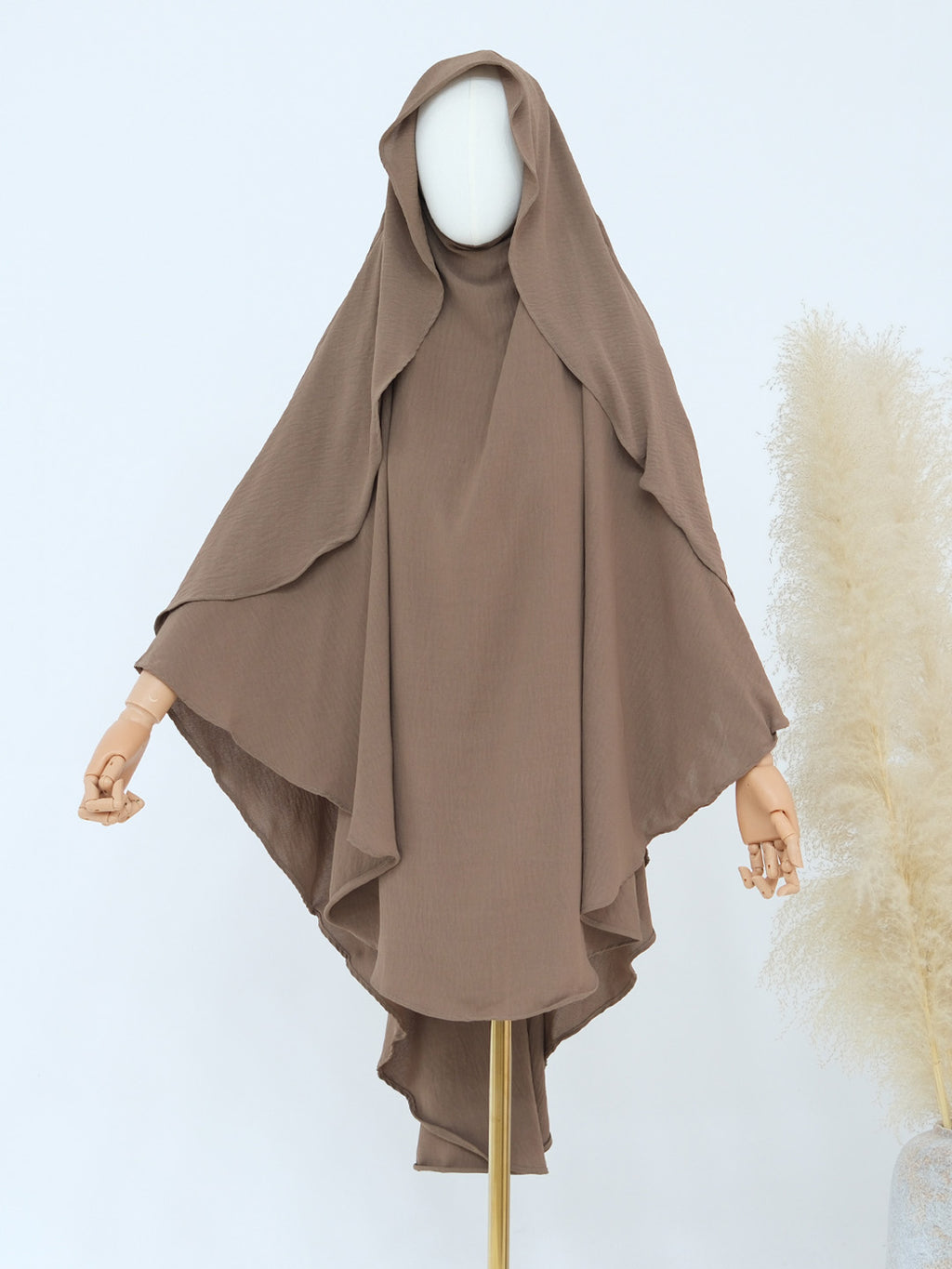 A taupe brown hijab with a draped hood and layered ruffle edges, displayed on a mannequin with wooden arms against a light blue background.