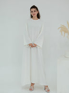 A woman modeling a long-sleeved white maxi dress with a round neckline and flowing skirt, paired with white heeled sandals, set against a clean white studio background.