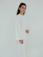 A woman modeling a long-sleeved white dress with a round neckline and flowing, wide sleeves, set against a plain white studio background.