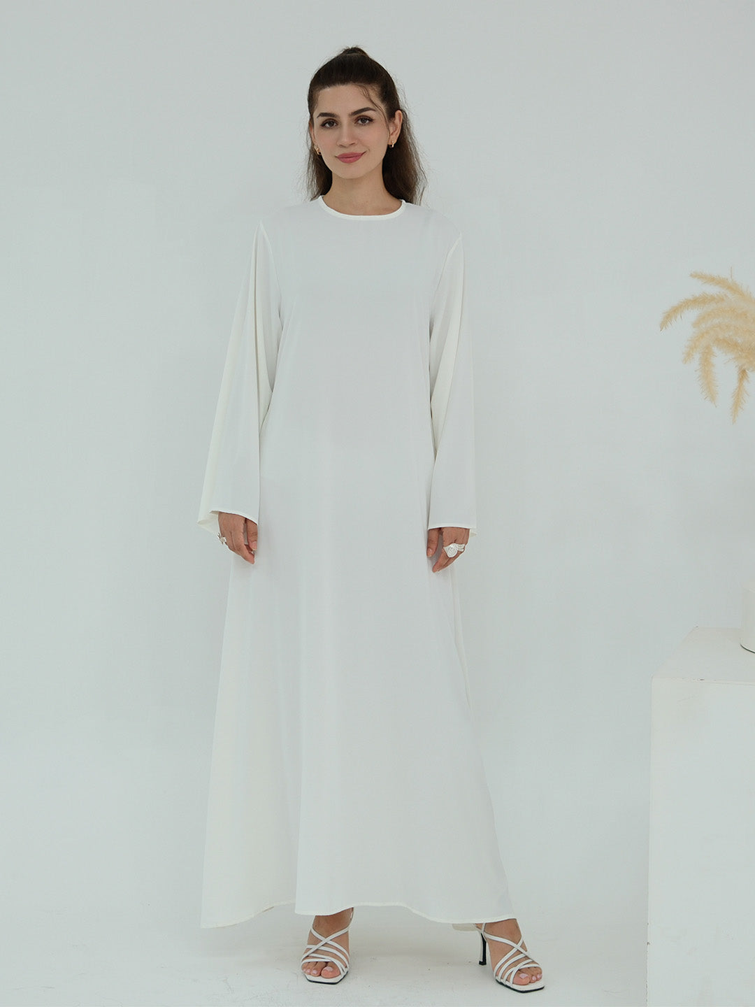 A woman modeling a long-sleeved white maxi dress with a round neckline, standing against a clean white studio background.
