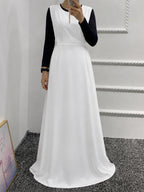 A woman wearing a long white dress with black long sleeves, taking a mirror selfie in an indoor setting with a white wall and gray carpet.