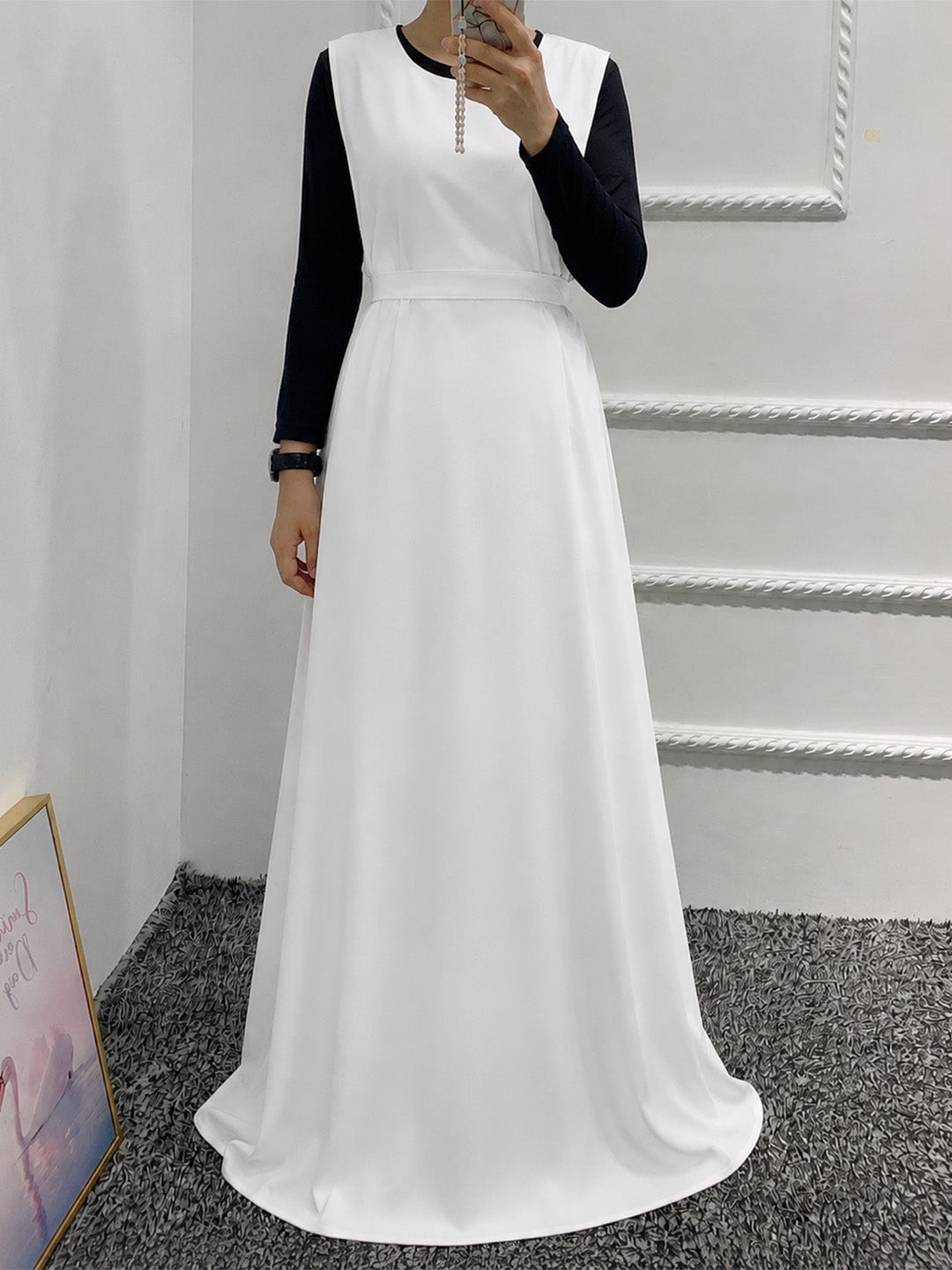 A woman wearing a long white dress with black long sleeves, taking a mirror selfie in an indoor setting with a white wall and gray carpet.