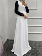 A woman wearing a sleeveless white maxi dress over a black long-sleeve top, taking a mirror selfie in an indoor setting with a gray carpet and white paneled walls.