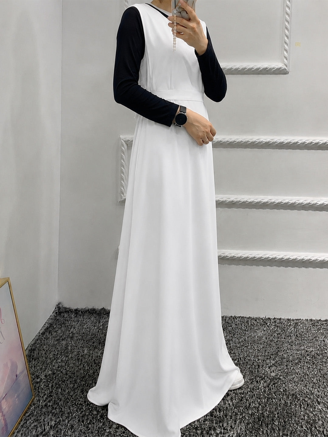 A woman wearing a sleeveless white maxi dress over a black long-sleeve top, taking a mirror selfie in an indoor setting with a gray carpet and white paneled walls.