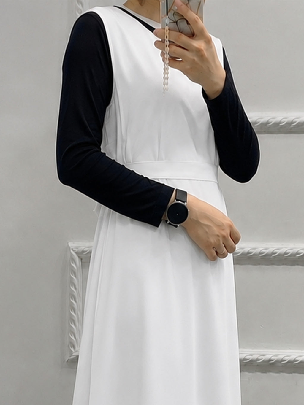 A person wearing a white sleeveless dress layered over a black long-sleeve top, holding a phone in a mirror selfie against a light gray wall with decorative molding.