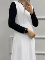 A person wearing a white sleeveless dress layered over a black long-sleeve top, holding a phone in a mirror selfie against a light gray wall with decorative molding.