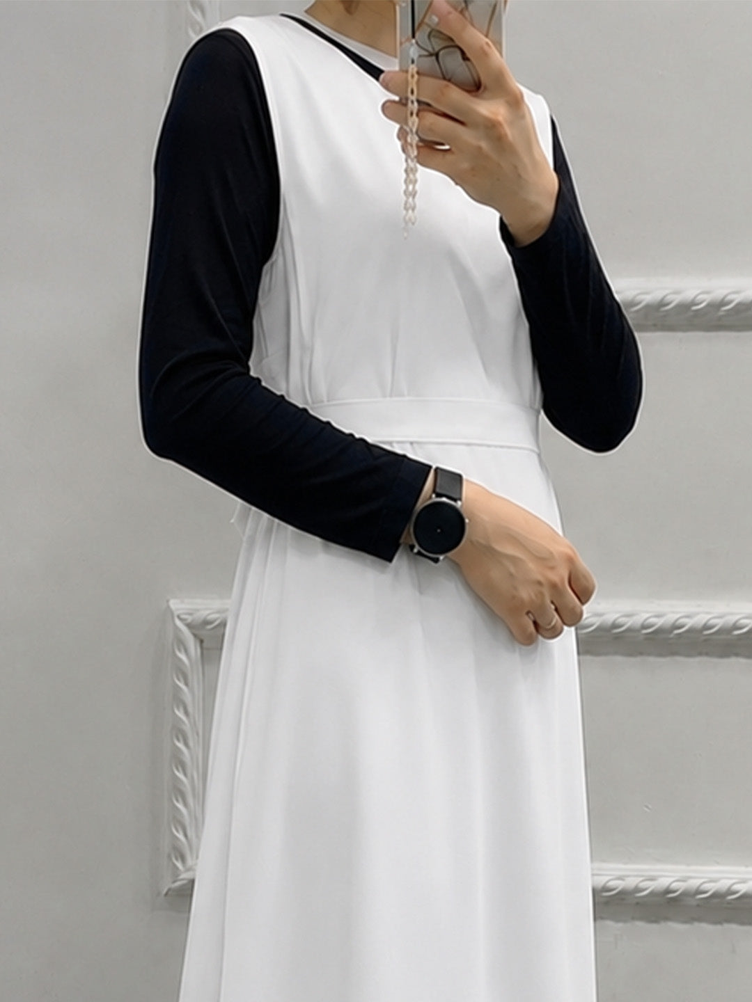 A person wearing a white sleeveless dress layered over a black long-sleeve top, holding a phone in a mirror selfie against a light gray wall with decorative molding.