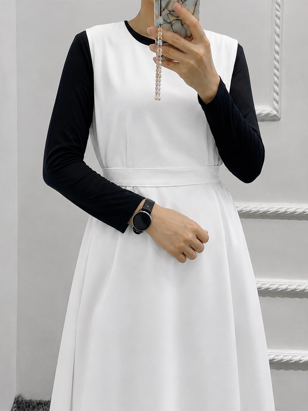 A woman wearing a white sleeveless dress layered over a black long-sleeve top, taking a mirror selfie. She is wearing a black smartwatch and holding a phone with a pearl necklace.