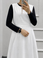 A woman wearing a white sleeveless dress layered over a black long-sleeve top, taking a mirror selfie. She is wearing a black smartwatch and holding a phone with a pearl necklace.