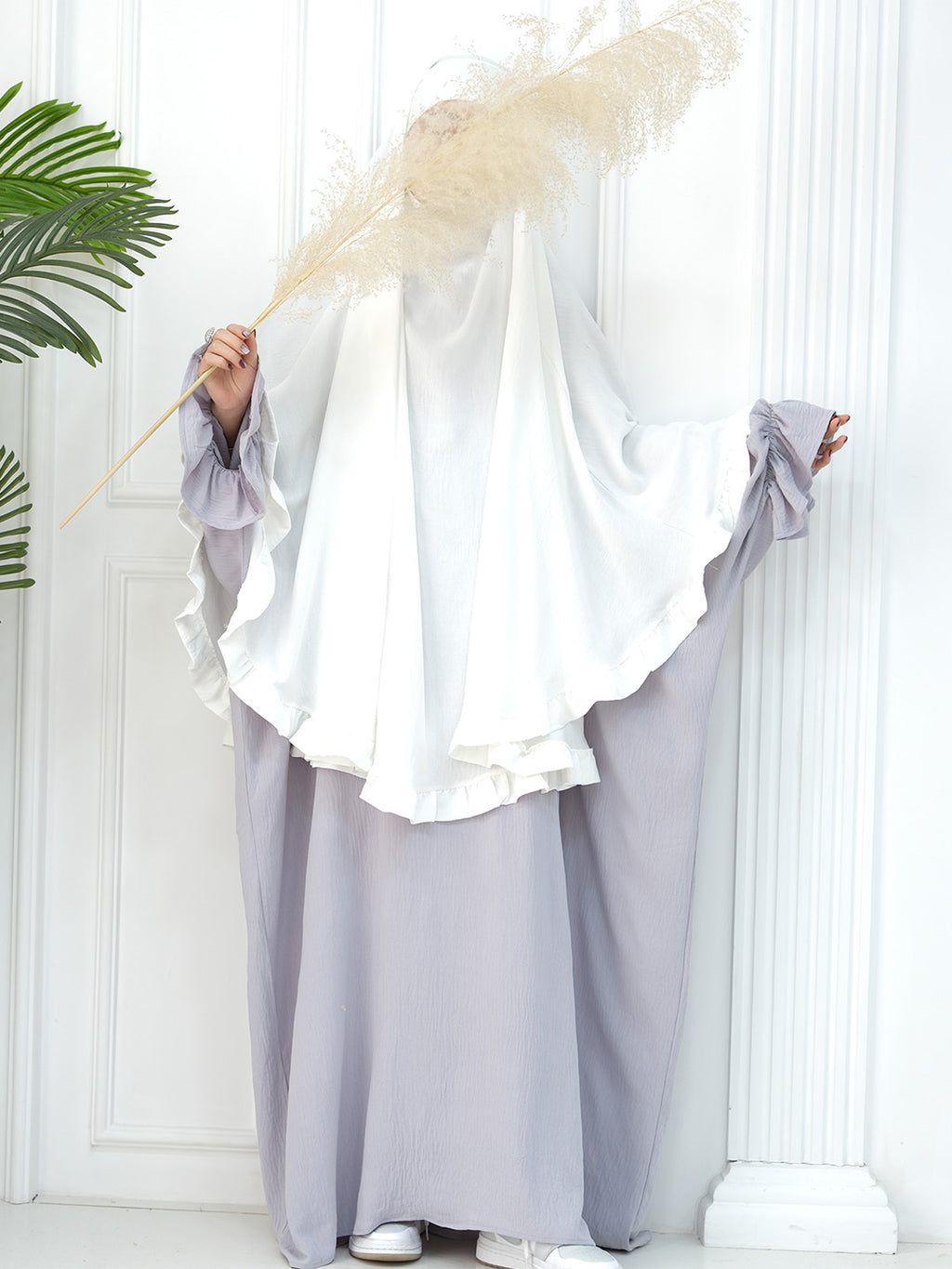 A person wearing a modest white and light gray/lavender abaya with a full-length head covering, holding a large pampas grass stem in front of a white paneled wall.