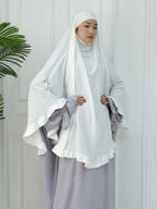 A woman wearing a white modest outfit with a large flowing hijab and wide flutter sleeves, paired with a light lavender skirt, standing against a white paneled wall with a green plant leaf visible on the left.