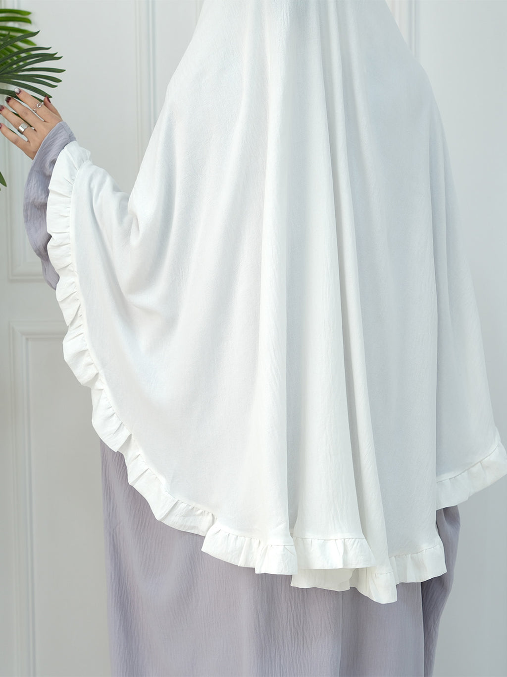 A close-up of a person wearing a flowing white modest garment with wide draped sleeves and ruffled trim, paired with a light lavender skirt. A hand with rings is visible holding a green plant leaf against a white paneled wall.