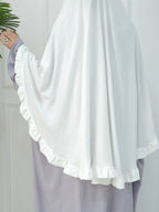 A close-up of a person wearing a flowing white modest garment with wide draped sleeves and ruffled trim, paired with a light lavender skirt. A hand with rings is visible holding a green plant leaf against a white paneled wall.