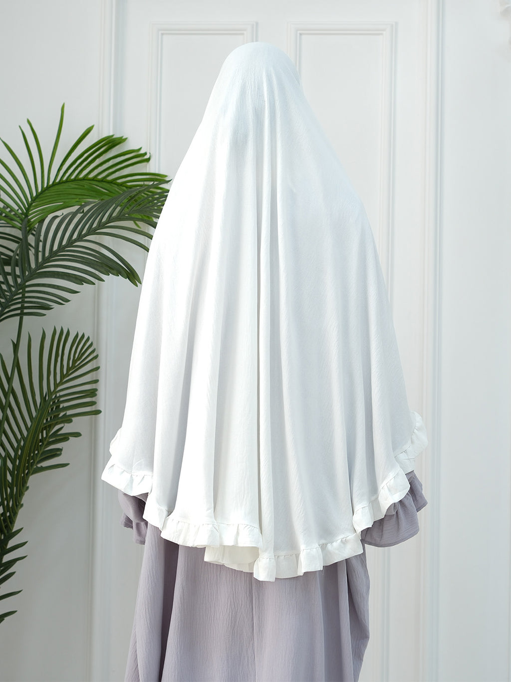A white modest headscarf or hijab draped over a person's head and shoulders, paired with a light lavender dress, standing in front of a white paneled door with a green plant on the left.