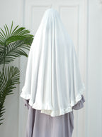 A white modest headscarf or hijab draped over a person's head and shoulders, paired with a light lavender dress, standing in front of a white paneled door with a green plant on the left.