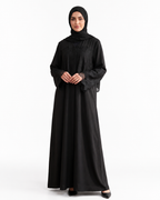 A woman modeling a full-length black abaya with a matching black hijab, standing against a plain white studio background.