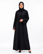 A woman modeling a black long-sleeve abaya with lace cuffs and a matching black hijab, standing against a plain white studio background.