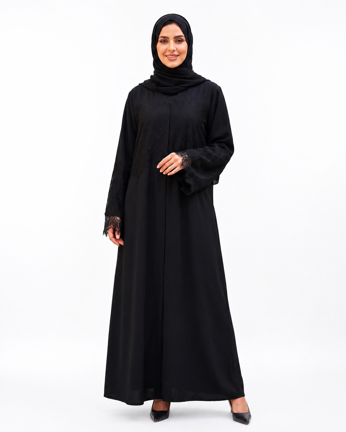 A woman modeling a black long-sleeve abaya with lace cuffs and a matching black hijab, standing against a plain white studio background.