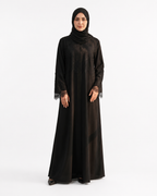 A woman modeling a long black abaya with matching black hijab, standing against a plain white studio background. The abaya has subtle tonal texture and lace trim at the cuffs.