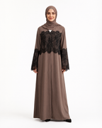 A woman modeling a taupe-brown long abaya dress with black lace panels on the chest and sleeves, paired with a matching hijab, against a plain white studio background.